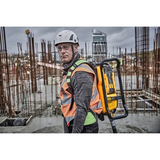 A construction worker wearing a helmet and safety vest is carrying a DEWALT DCPS310N photography equipment on their back at a construction site with metal rebar structures and concrete flooring visible in the background.