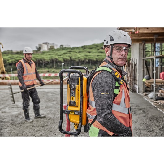 A construction worker wearing a safety vest and helmet is carrying a DEWALT DCPS310N power tool on their back at an outdoor construction site, with another worker seen in the background holding a rake. Faces are blurred for privacy.