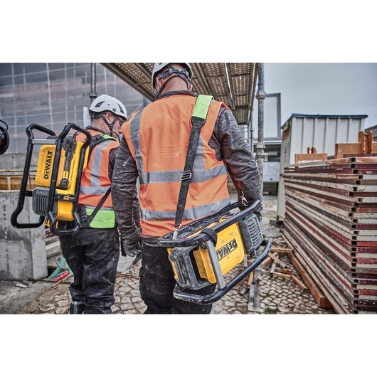 Two construction workers wearing safety helmets and orange reflective vests are walking on a construction site, each carrying a portable DEWALT power tool.