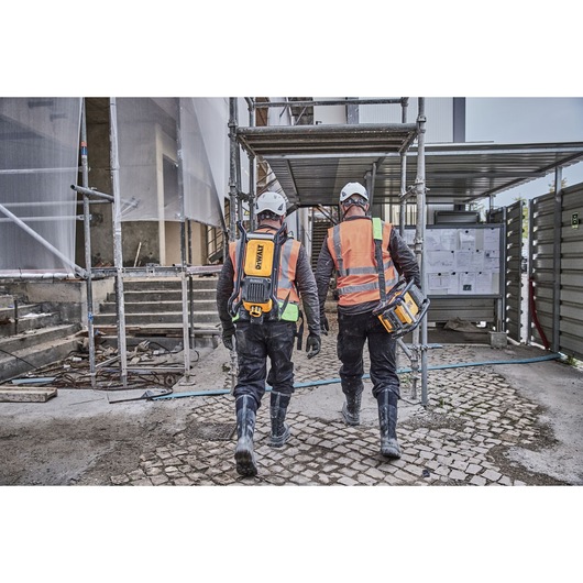 Two construction workers wearing safety helmets and orange reflective vests are walking at a construction site. One worker carries a DEWALT power tool and the other wears a DEWALT backpack. Scaffolding and building materials are visible in the background.