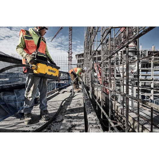 Two construction workers in safety vests operate a DEWALT power tool on a building site. One worker is holding the yellow DEWALT device, while the other is working near steel reinforcement bars. The scene includes industrial structures, cranes, and building materials.