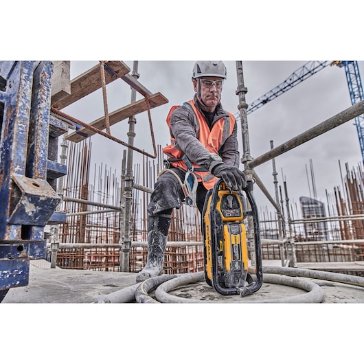 A construction worker wearing safety gear and an orange vest uses a DEWALT DCPS320N tool at an active construction site with scaffolding and building materials visible in the background.
