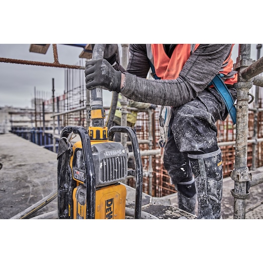 A construction worker using a DEWALT power tool, surrounded by scaffolding and wearing protective gear. The tool is in operation on a building site with visible concrete and rebar structures.
