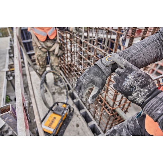 A close-up of a construction worker wearing gloves and pressing a button on their wrist device. They are standing next to a metal rebar structure with a DEWALT tool on the ground. Another worker is visible in the background, both wearing safety gear.