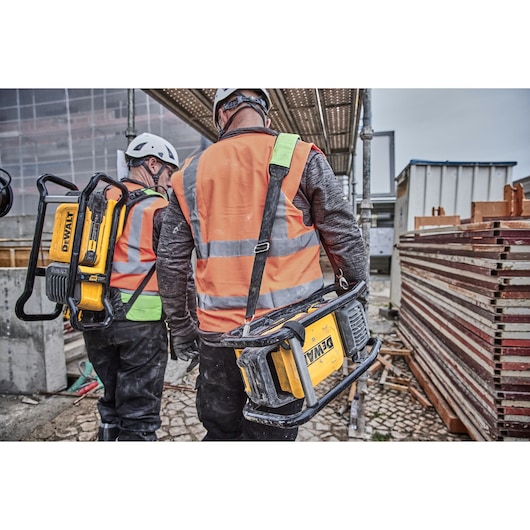 Two workers wearing orange reflective vests and helmets are walking on a construction site, each carrying a portable DEWALT device with sturdy handles.