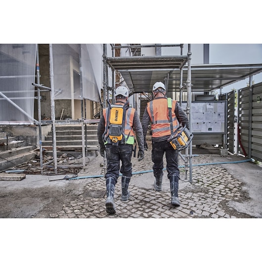 Two construction workers wearing safety helmets, orange vests, and boots walk on a building site. Each is carrying DEWALT equipment, with one worker wearing a backpack-style device and the other holding a portable tool. Scaffolding and construction materials are visible around them.