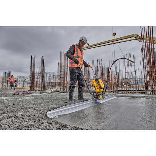 A construction worker wearing an orange safety vest and helmet operates a DEWALT power tool to smooth freshly poured concrete at a building site with exposed steel rods and construction equipment under a cloudy sky.