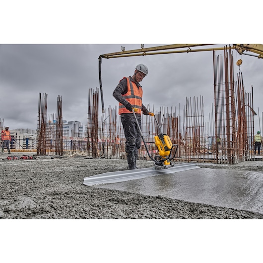A construction worker wearing a helmet and orange safety vest uses a DEWALT concrete screed tool on a building site with steel reinforcement bars and wet concrete.