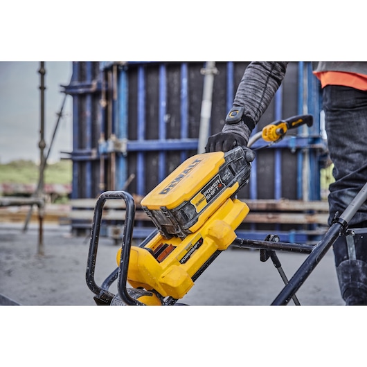 A close-up view of a worker using a DEWALT Power Shift battery-powered tool on a construction site. The yellow device is held by a person wearing gloves and work clothing, with concrete and scaffolding visible in the background.