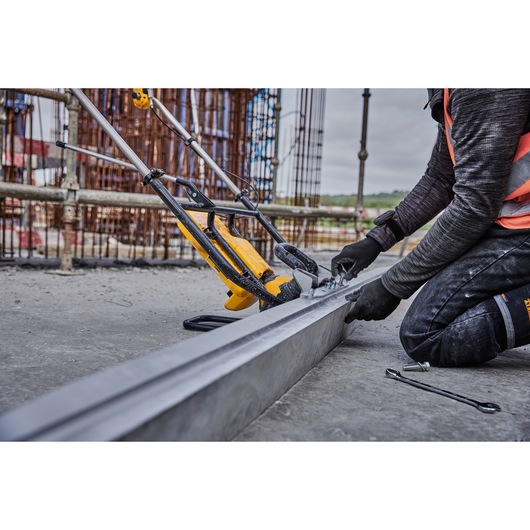 A person wearing work gloves and safety gear is assembling or adjusting a DEWALT power tool on a construction site. The tool is positioned alongside a metal beam, with other construction equipment and scaffolding visible in the background.