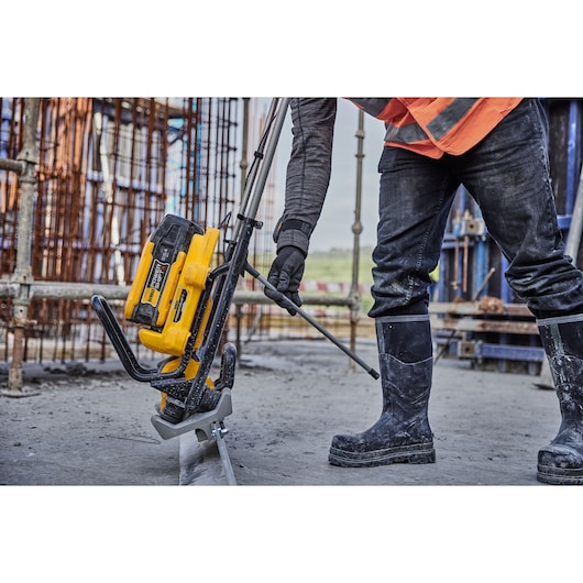 A DEWALT cordless tool (DCPS330N) is being used by a worker wearing rubber boots and safety gear on a construction site. The worker is holding a long pole attached to the tool, which is positioned on the ground near metal rebar and scaffolding.