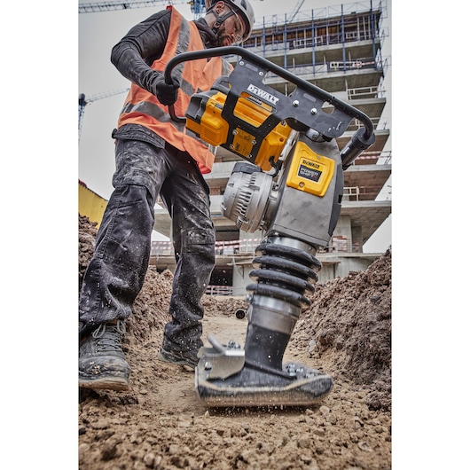 A person wearing safety gear operates a DEWALT DCPS660N plate compactor on a construction site, compacting soil with a building structure visible in the background.