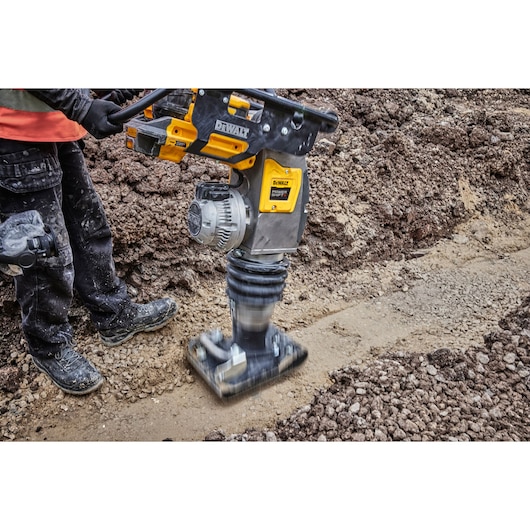 A DEWALT battery-powered compactor tool (DCPS660N) being operated by a person on a construction site, compacting soil in a trench. The tool features yellow DEWALT branding and is designed for professional earthwork applications.