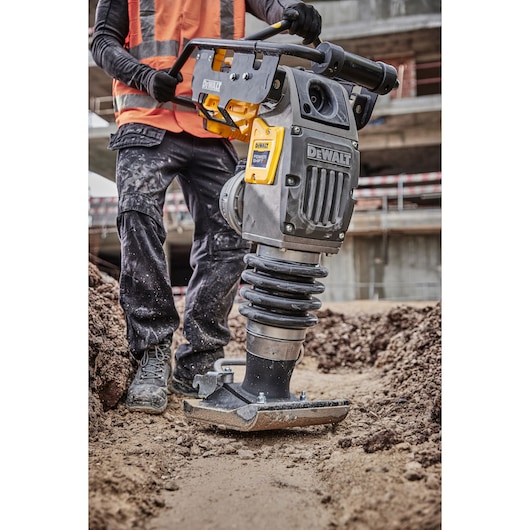 A person in work clothes and a safety vest operates a DEWALT-branded compactor machine, tamping soil at a construction site.