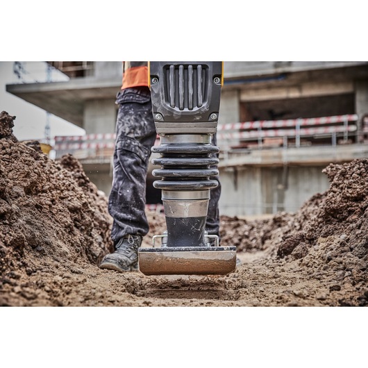 Close-up of a DEWALT tool being used to compact soil at a construction site. The lower part of a worker is visible, standing in the dirt with the machine. The background shows building structures and barriers.