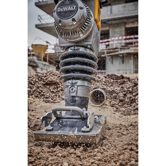A close-up view of a DEWALT compactor tool in action on a construction site, compacting soil with dust particles flying around.