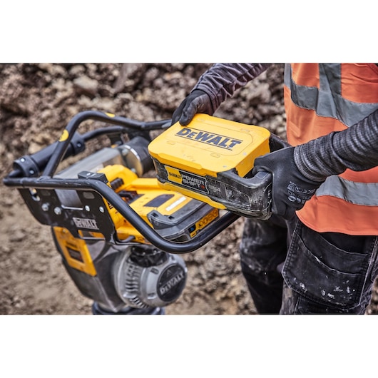 A person wearing gloves and a high-visibility vest is holding a DEWALT Power Shift battery near a yellow DEWALT tool or machine on a construction site.