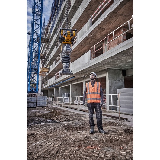 A person wearing a safety helmet and orange high-visibility vest stands on a construction site near a building under construction. Above them, a DEWALT DCPS660N tool is suspended in the air, likely being moved with construction machinery.