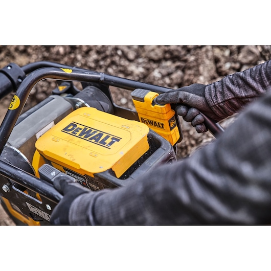 Close-up of a DEWALT outdoor power tool with a yellow casing, showing a gloved hand operating the controls. The tool is being used on a rugged, earthy surface.