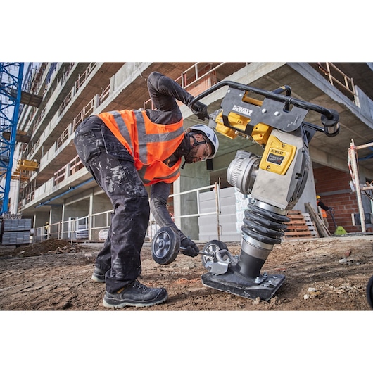 A construction worker wearing a safety helmet and orange reflective vest is using a DEWALT power tool on a construction site. The worker is operating the DEWALT tool on bare ground near a large building under construction.