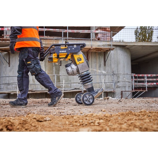 A person in workwear and a high-visibility vest is pulling a DEWALT tool with wheels, on a construction site with building structures and scaffolding in the background.
