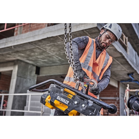 A construction worker wearing a helmet and high-visibility vest is handling a DEWALT DCPS660N tool using a lifting chain at a building site. The tool is prominently marked with the DEWALT logo, and the worker's face is blurred for privacy.