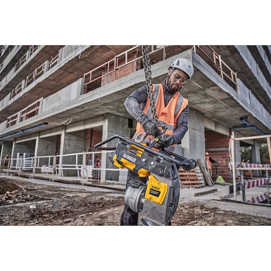 A person wearing safety gear uses a DEWALT DCPS660N power tool at a construction site in front of a partially built concrete building.