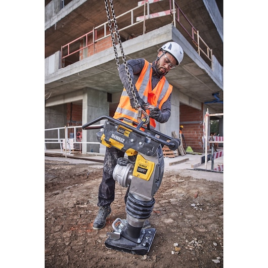 A construction worker wearing a safety helmet and orange reflective vest is operating a DEWALT DCPS660N compactor machine at a building site. The worker is attaching a chain to the machine on a dirt ground with a partially constructed building in the background.