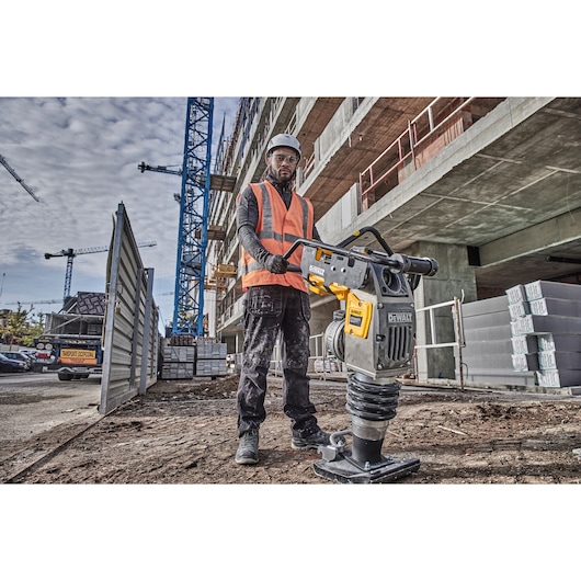 A construction worker wearing a safety vest and helmet operates a DEWALT DCPS660N power tool on a construction site, with a partially constructed building and cranes visible in the background.