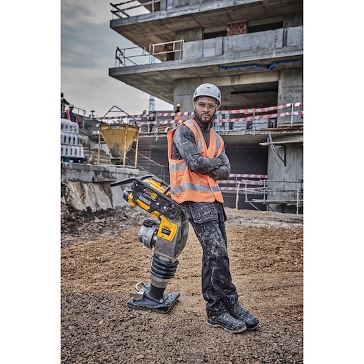 A construction worker wearing a safety helmet and orange high-visibility vest stands with arms crossed next to a DEWALT DCPS660N power tool on a construction site with a partially built building in the background.