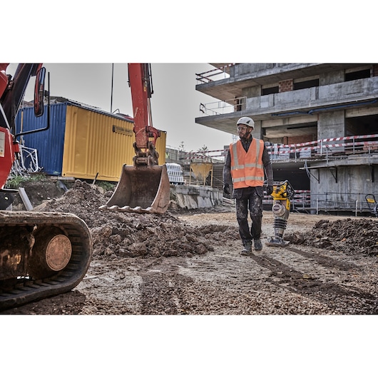 A construction worker wearing a safety helmet and orange high-visibility vest operates a yellow DEWALT power tool on a dirt surface at a construction site. An excavator and a yellow shipping container are visible in the background, along with a partially built concrete structure.