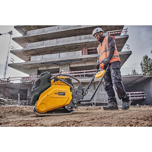 A construction worker wearing a helmet and safety vest operates a yellow DEWALT plate compactor on a dirt surface at a building site, with a multi-story concrete structure in the background.