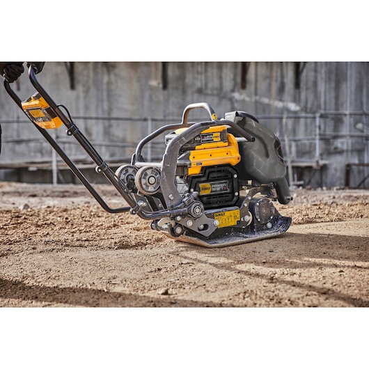 A DEWALT plate compactor machine operating on a construction site, compacting soil. The equipment features a sturdy metal frame, yellow DEWALT branding, and a visible battery compartment.