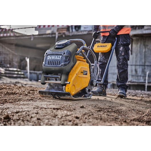 A close-up view of a DEWALT plate compactor being operated on a construction site by a worker wearing dark pants and a high-visibility vest.