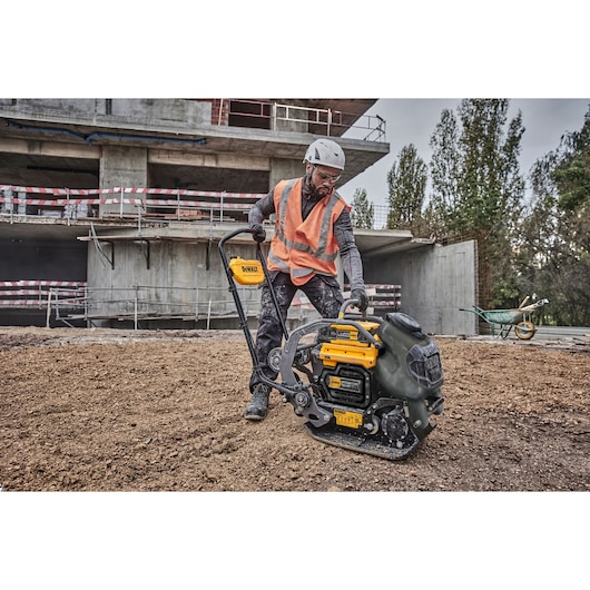 A construction worker wearing a helmet and orange safety vest operates a DEWALT DCPS7154N plate compactor on a building site with a partially constructed structure and trees in the background.
