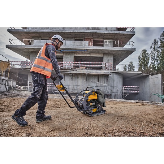A construction worker wearing a safety helmet and high-visibility vest operates a DEWALT plate compactor on a gravel surface at a building site, with a partially constructed concrete structure in the background.