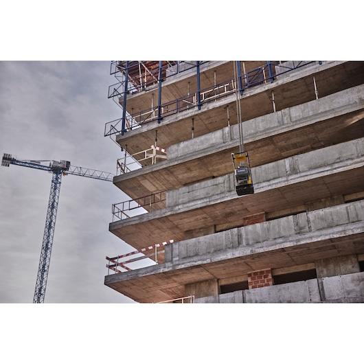 A DEWALT power tool, model DCPS7154N, is being hoisted on a chain alongside a multi-story concrete building under construction. A tower crane is visible on the left side against a cloudy sky.
