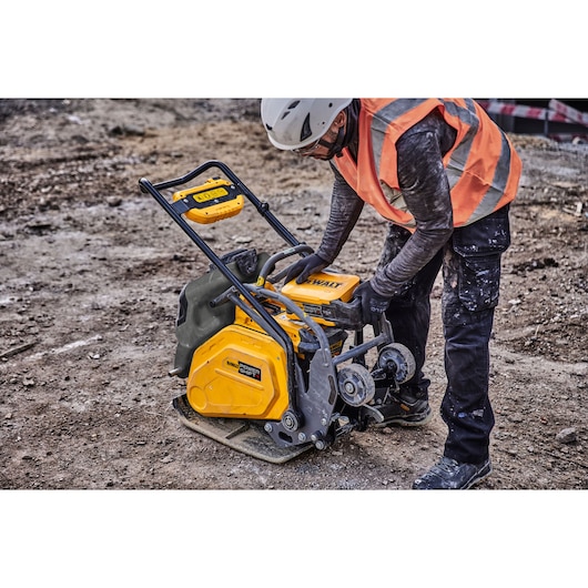 A construction worker wearing a helmet and orange safety vest operates a DEWALT plate compactor on a dirt surface at a construction site.