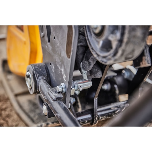 Close-up of a hand wearing a glove adjusting a metallic component on a DEWALT power tool, showing bolts and metal parts with a yellow casing visible in the background.