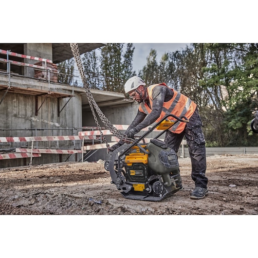 A construction worker wearing a safety helmet and orange vest is handling a DEWALT plate compactor (SKU: DCPS7154N) on a building site. The worker is attaching chains to the machine, which is positioned on uneven soil near a partially constructed concrete structure.