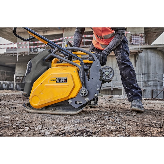 A DEWALT Power Shift plate compactor, model DCPS7154N, is being operated by a construction worker on a dirt surface at an outdoor job site, with concrete structures visible in the background.
