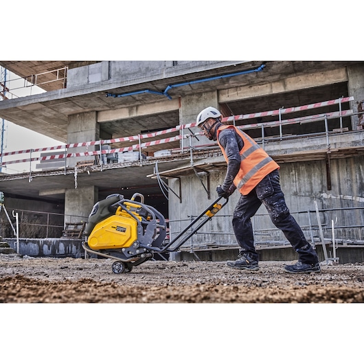A construction worker wearing a helmet and an orange safety vest operates a yellow DEWALT plate compactor on a dirt construction site in front of a partially built concrete structure.