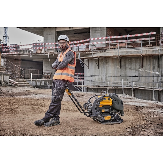 A construction worker wearing a helmet and safety vest stands next to a DEWALT plate compactor on a dirt surface at a construction site. The worker's face is blurred, and a partially built structure is visible in the background.