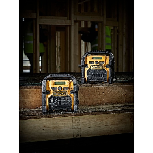 Two DEWALT DCR019 portable radios are placed on wooden beams in a construction site. The radios are yellow and black with a rugged design, showing dust and wear from use.