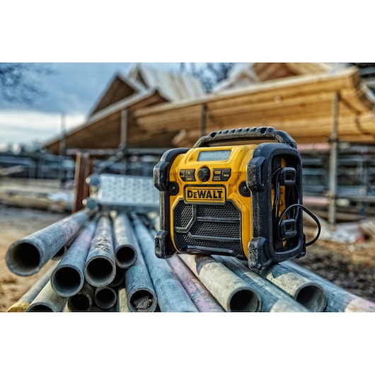A yellow and black DEWALT portable radio, model DCR020, is placed on several metal pipes at a construction site with wooden