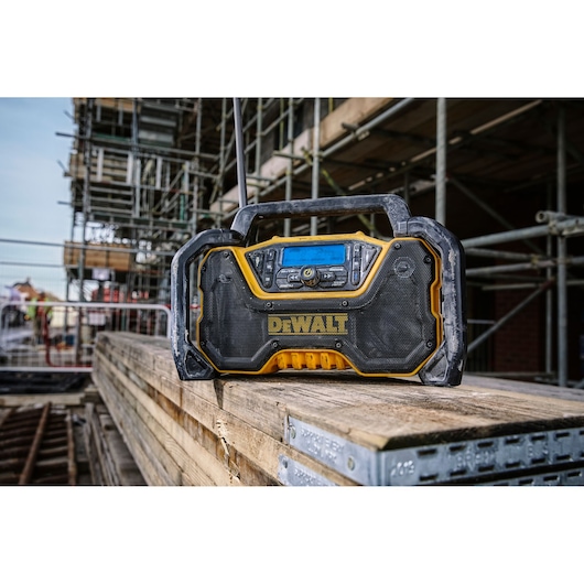A DEWALT portable radio with rugged design sits on a stack of wooden planks at a construction site, with scaffolding and
