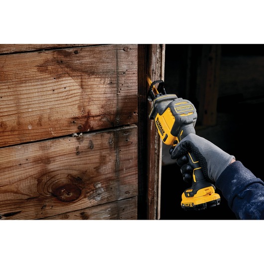 Gloved hand using a DEWALT cordless reciprocating saw on a wooden surface indoors.