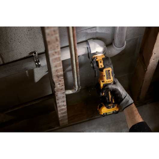 A person wearing a glove uses a DEWALT cordless oscillating multi-tool to cut a white PVC pipe near a wall and other pipes in a construction or repair setting.