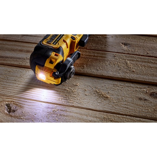 A DEWALT cordless oscillating multi-tool with a built-in LED light is shown in action on a wooden surface with wood dust around.