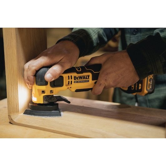 Person using a DEWALT cordless multi-tool to sand wood furniture corner indoors.